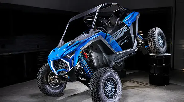 A front-angled shot of a Polaris RZR Pro R Ultra Edition Velocity Blue with one tire propped on a barrel to highlight its FOX suspension.