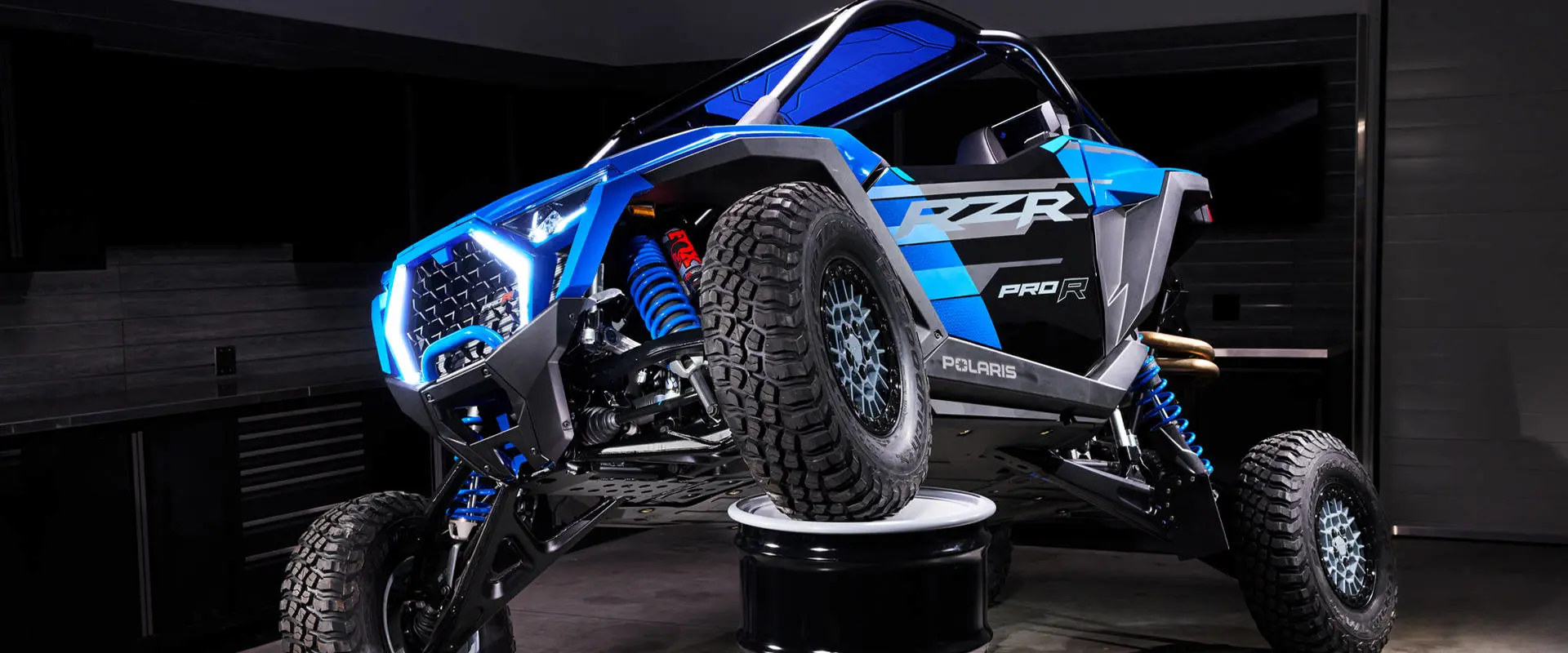 Side view of the RZR Pro R Ultra Edition Velocity Blue with a front wheel propped on a barrel to show off its FOX suspension.