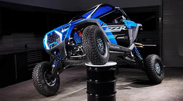 Side view of the RZR Pro R Ultra Edition Velocity Blue with a front wheel propped on a barrel to show off its FOX suspension.