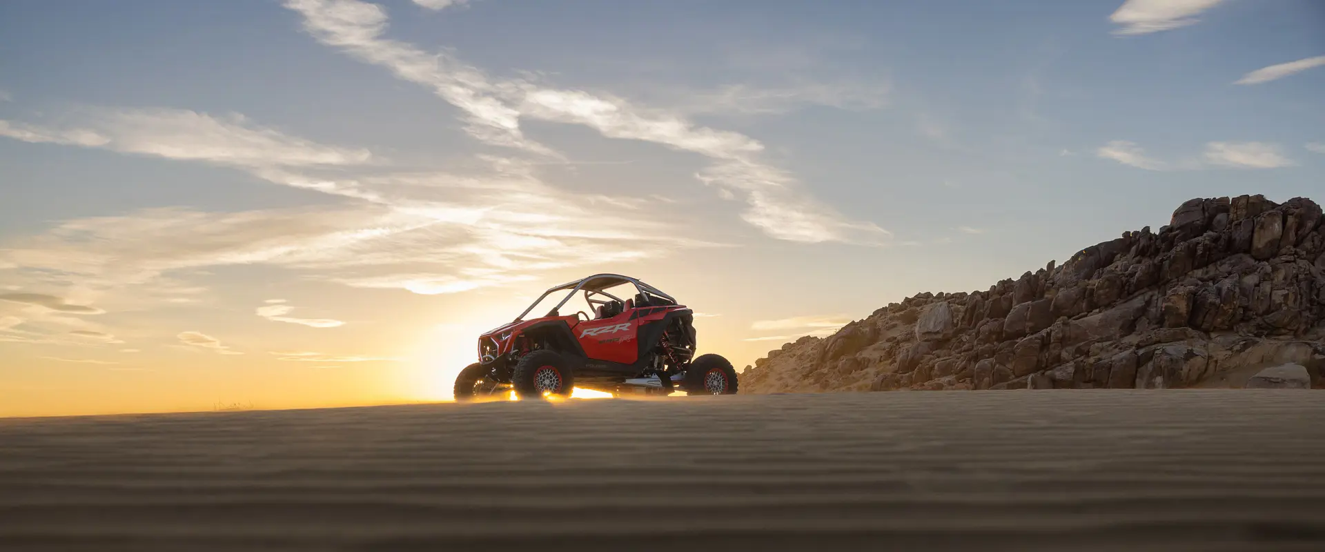 A 2026 Polaris RZR Pro R SxS in the Indy Red colorway parked on a hill in the desert as sunlight surrounds it.