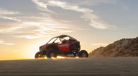A 2026 Polaris RZR Pro R SxS in the Indy Red colorway parked on a hill in the desert as sunlight surrounds it.