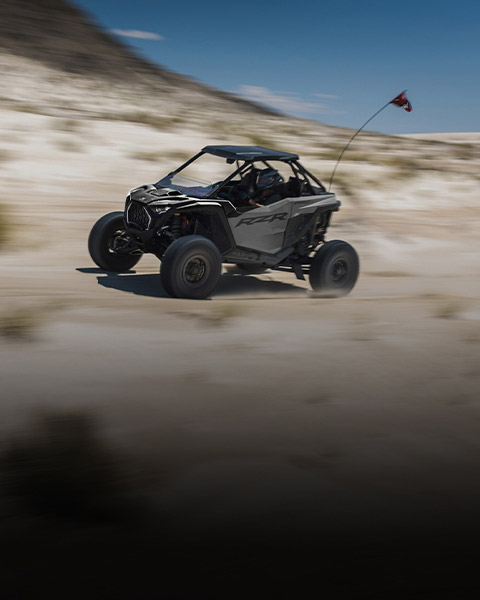 A gray Polaris RZR PRO S kicks up sand while speeding across desert dunes under a clear sky.