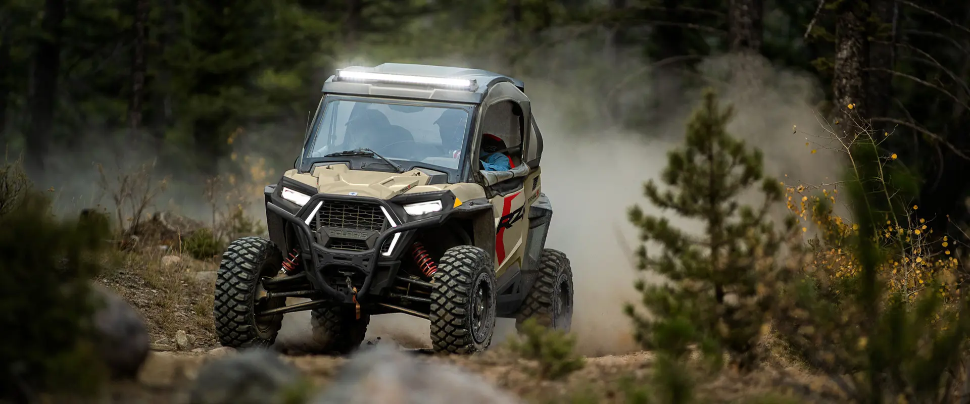 A 2026 Polaris RZR Trail S side by side with headlights and lightbar illuminated while driving on a trail.