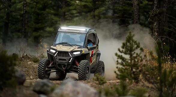 A 2026 Polaris RZR Trail S side by side with headlights and lightbar illuminated while driving on a trail.