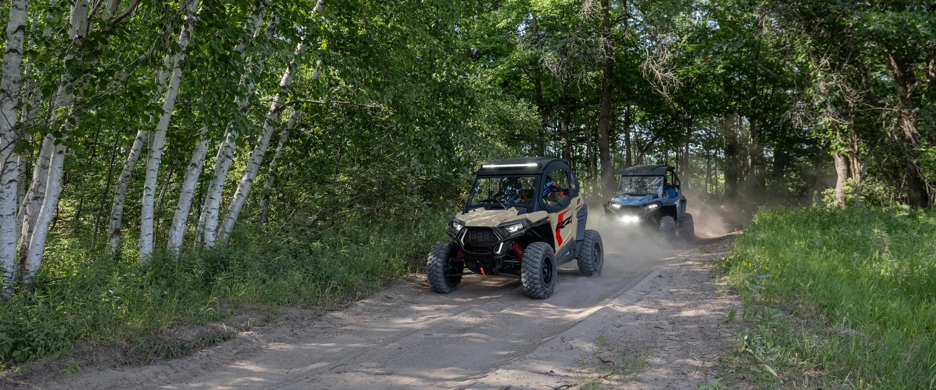 2 2026 Polaris RZR Trail side by sides cruising on a dusty forest trail.