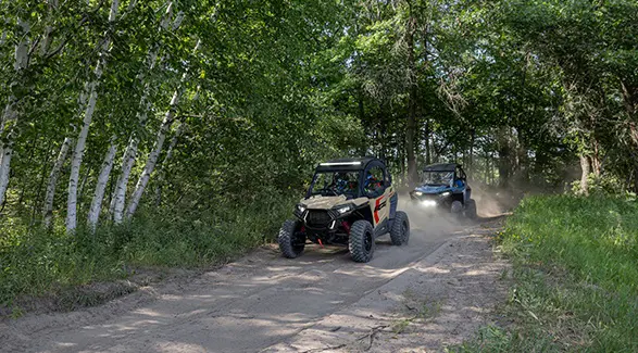 2 2026 Polaris RZR Trail side by sides cruising on a dusty forest trail.