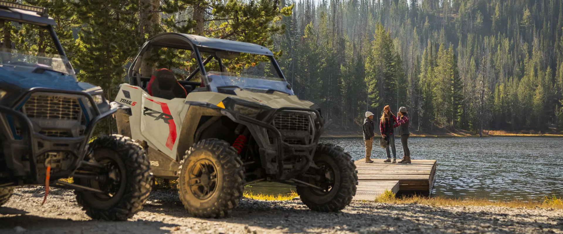 Closeup of a 2026 Polaris RZR Trail S side by side with 3 people in the background on a dock near a lake.