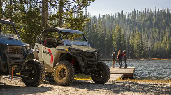 Closeup of a 2026 Polaris RZR Trail S side by side with 3 people in the background on a dock near a lake.