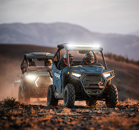 2 riders in a 2026 Polaris RZR Trail side-by-side with headlights and lightbar illuminated while cruising a trail.