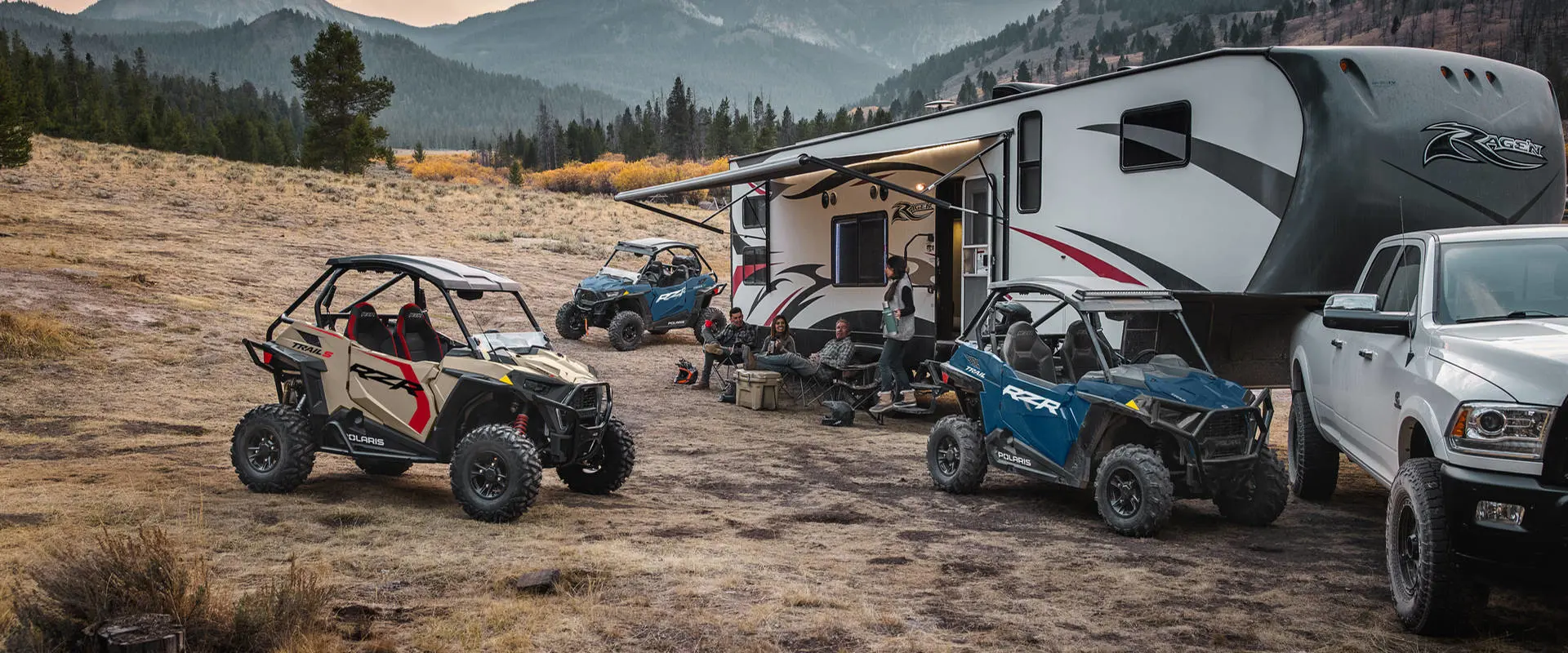 People relaxing near a camper while their 3 2026 Polaris RZR Trail side-by-sides are parked around them.