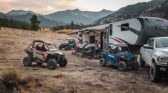 People relaxing near a camper while their 3 2026 Polaris RZR Trail side-by-sides are parked around them.