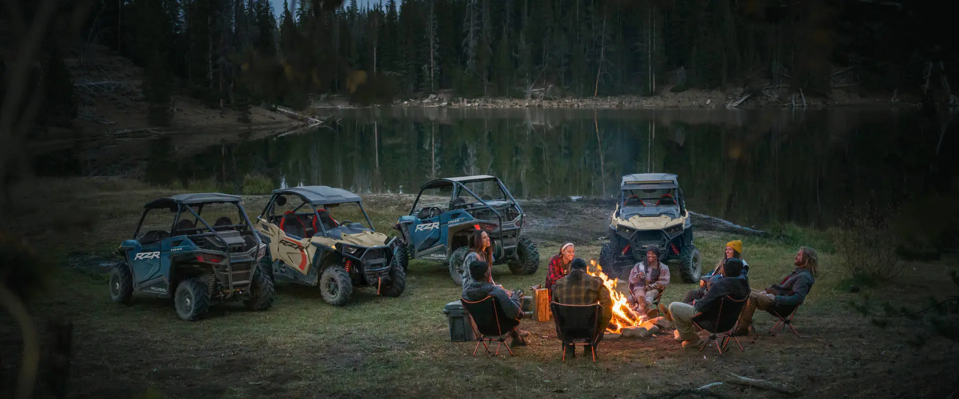 A group of friends around a campfire at dusk with their 4 2026 Polaris RZR Trail side-by-sides parked nearby.