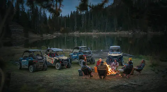 A group of friends around a campfire at dusk with their 4 2026 Polaris RZR Trail side-by-sides parked nearby.
