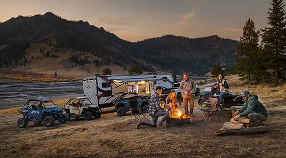 Friends around a campfire relaxing after a day of riding their 2026 Polaris RZR Trail side-by-sides.