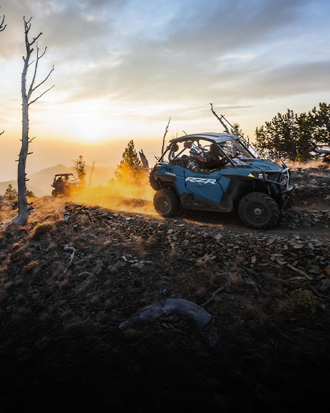 A blue Polaris RZR Trail vehicle drives on a dusty mountain path at sunset, with another RZR in the distance.
