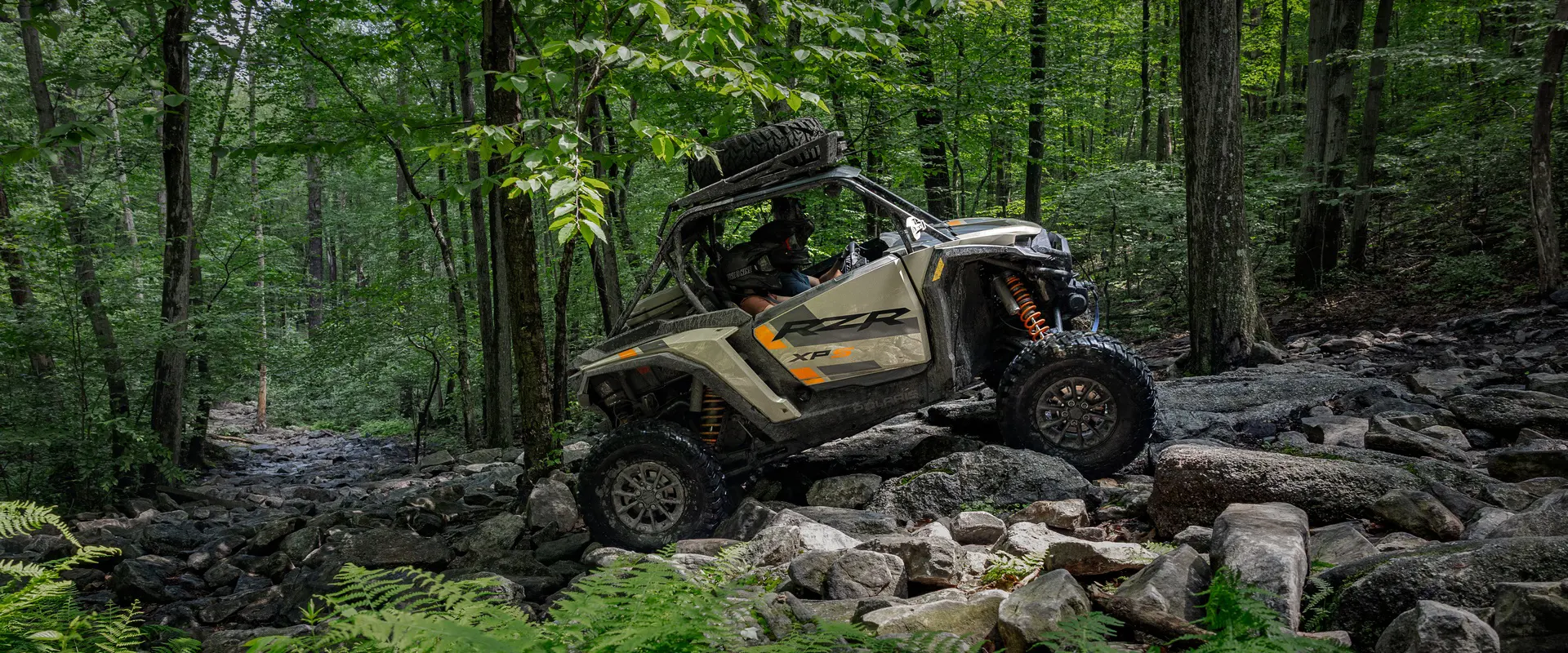 Side profile of a 2026 Polaris RZR XP S side-by-side crawling over rocky terrain in the forest.