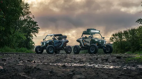 Two 2026 Polaris RZR XP S side-by-sides parked in an open muddy area with forest surrounding them.