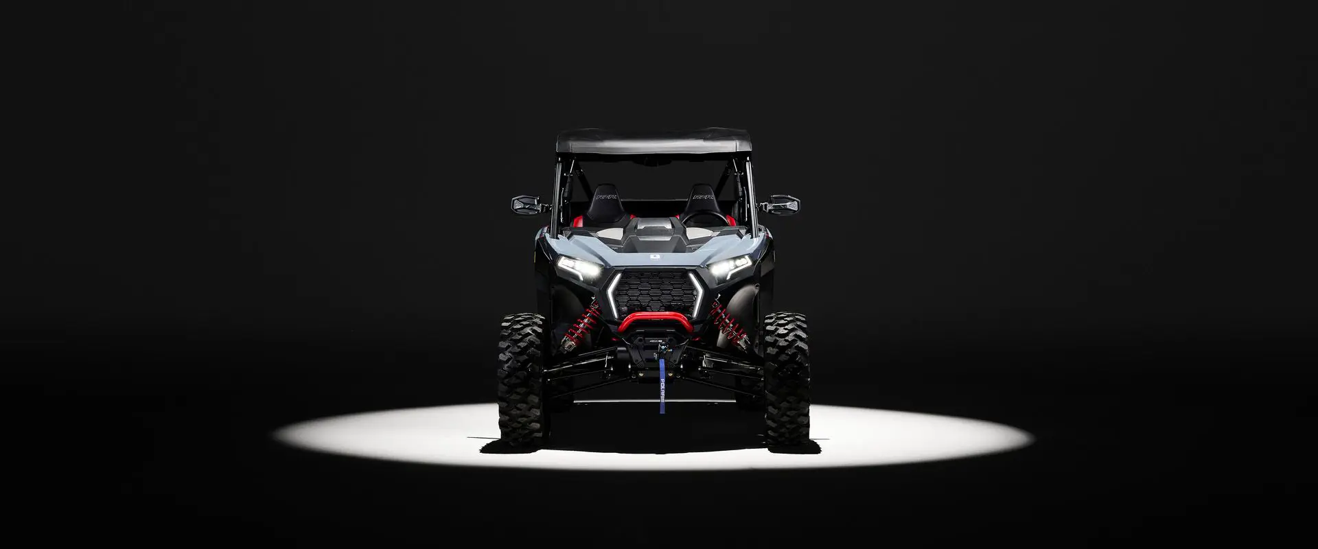 Front end view of a 2026 Polaris RZR XP Ultimate SxS in the Stealth Gray colorway entrenched in light while parked in a dark garage.