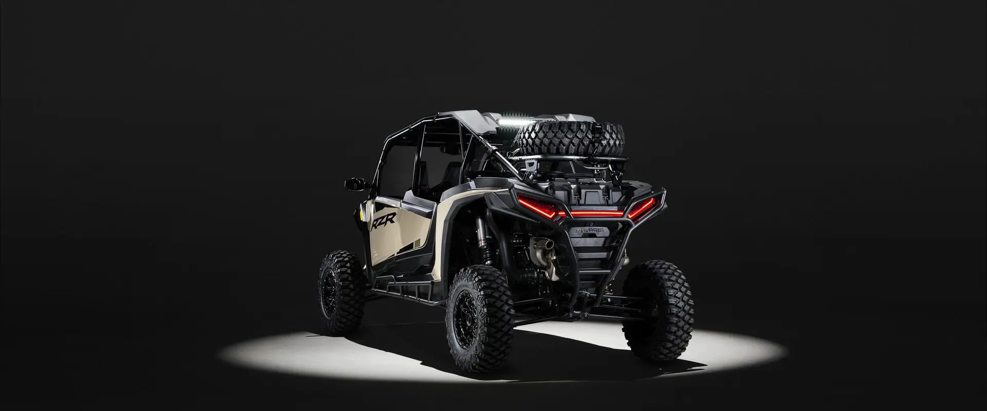 Rear view of a 2026 Polaris RZR XP 4 Ultimate SxS in the Sand Dune colorway entrenched in light while parked in a dark garage.