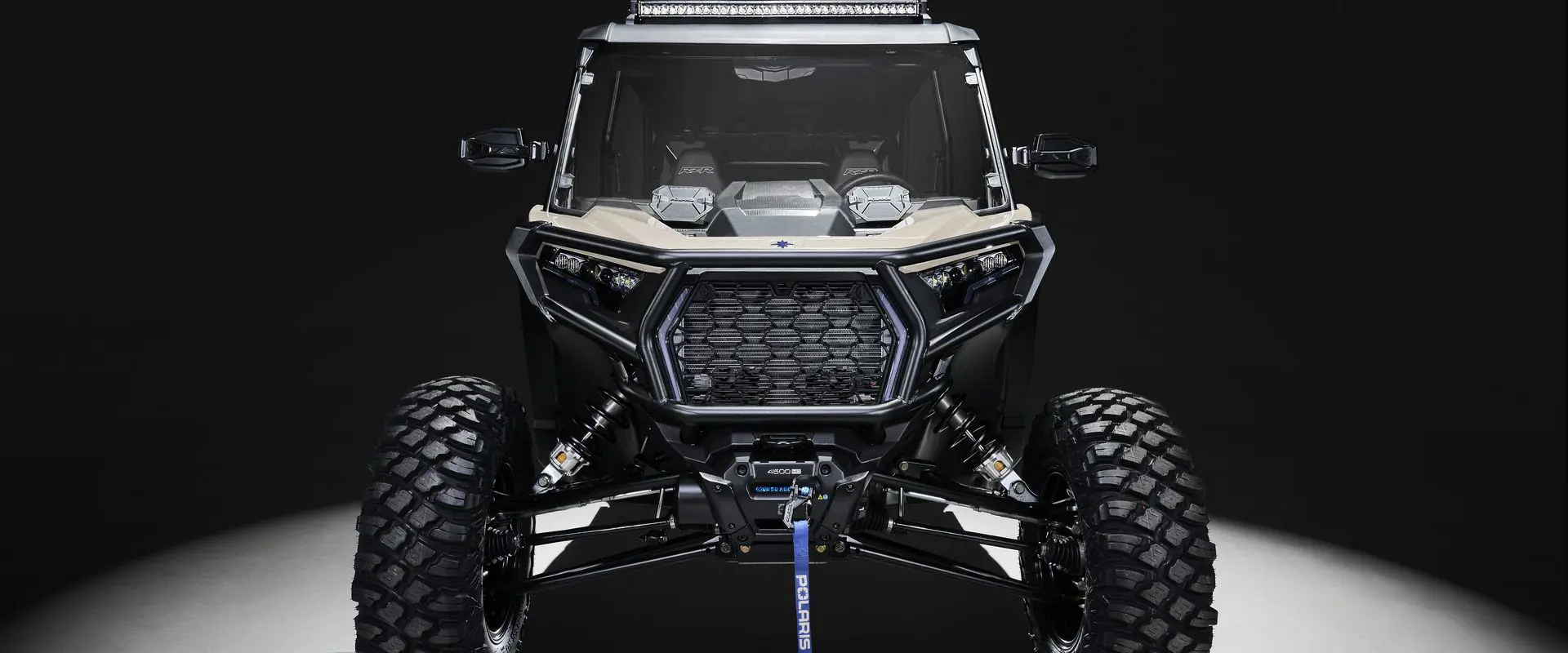 Front end view of a 2026 Polaris RZR XP 4 Ultimate SxS in the Sand Dune colorway entrenched in light while parked in a dark garage.