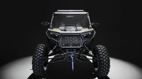 Front end view of a 2026 Polaris RZR XP 4 Ultimate SxS in the Sand Dune colorway entrenched in light while parked in a dark garage.