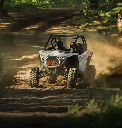 2 riders in a Polaris RZR sport side-by-side kicking up dirt on a sandy trail in the forest.