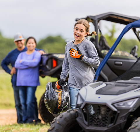 A 10 year old kid running toward a 2027 Polaris RZR 200 EFI youth side-by-side while her parents smile in the background.