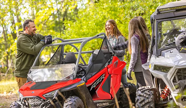 3 girls and a man chatting around a 2027 Polaris RZR 200 EFI youth side-by-side.