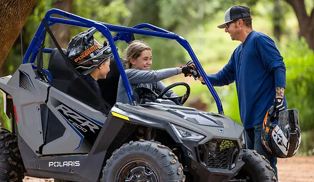 2 kids smiling at their dad while sitting in a 2027 Polaris RZR 200 EFI youth side-by-side in the Storm Gray colorway.