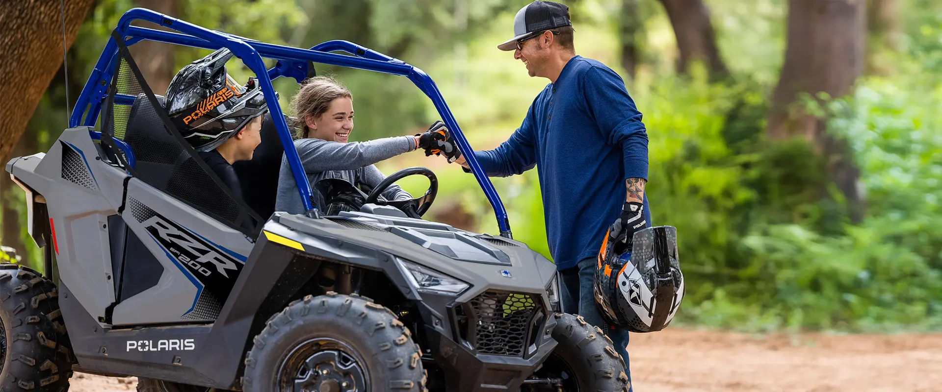 2 kids smiling at their dad while sitting in a 2027 Polaris RZR 200 EFI youth side-by-side in the Storm Gray colorway.