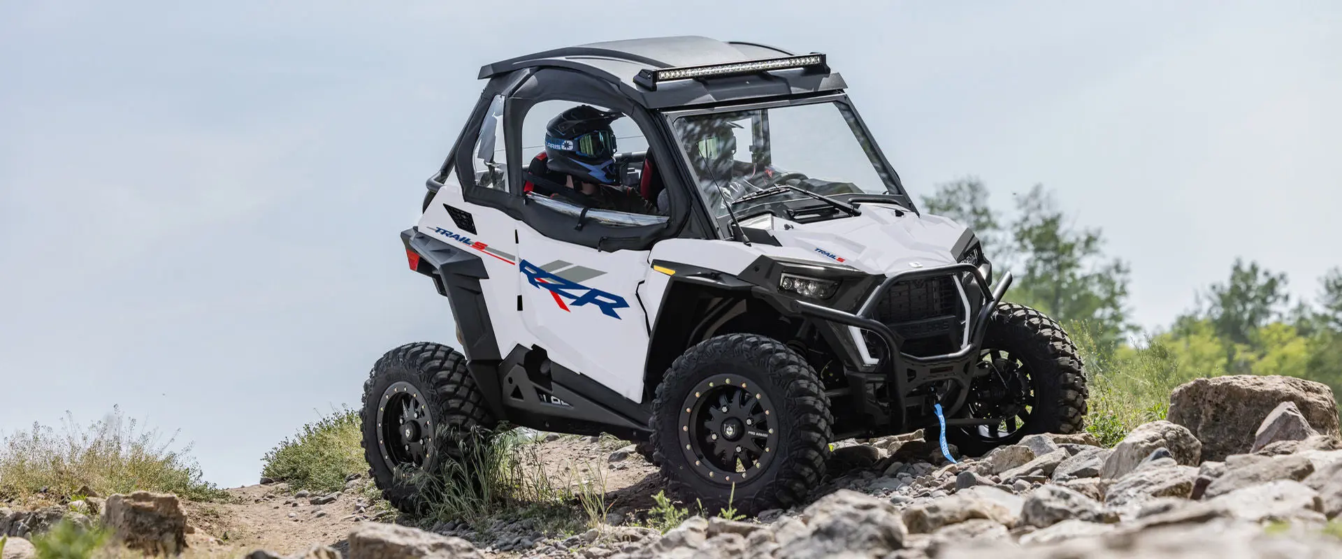 2 riders in a 2027 Polaris RZR Trail S side-by-side crawling over rocky terrain.