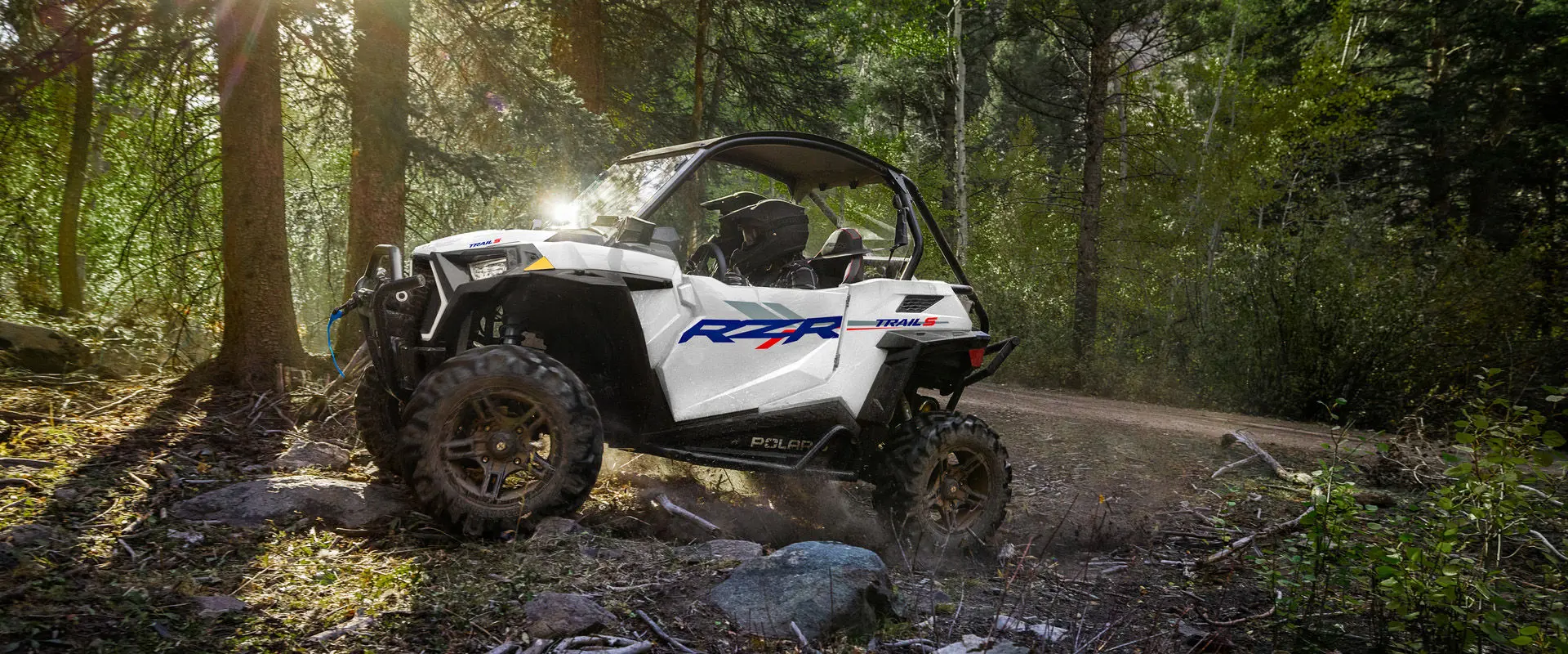 Side profile of a 2027 Polaris RZR Trail S side-by-side driving through the forest.
