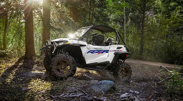 Side profile of a 2027 Polaris RZR Trail S side-by-side driving through the forest.