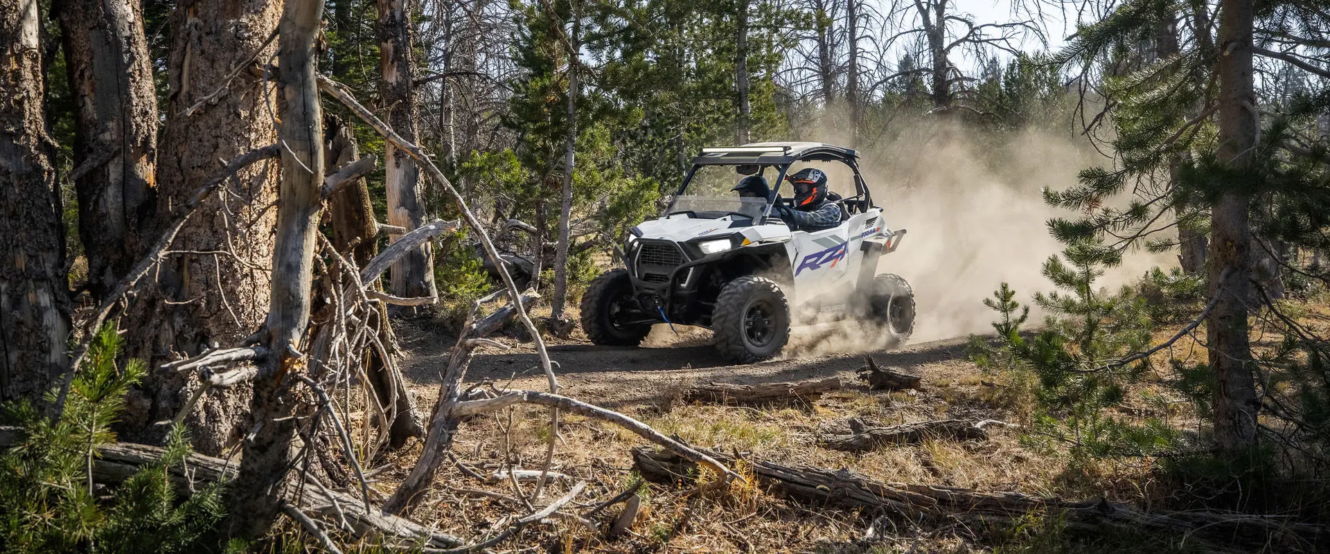 2 riders in a 2027 Polaris RZR Trail S side-by-side kicking up dust in the forest.