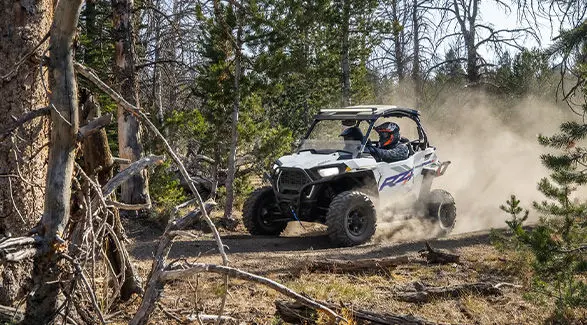 2 riders in a 2027 Polaris RZR Trail S side-by-side kicking up dust in the forest.
