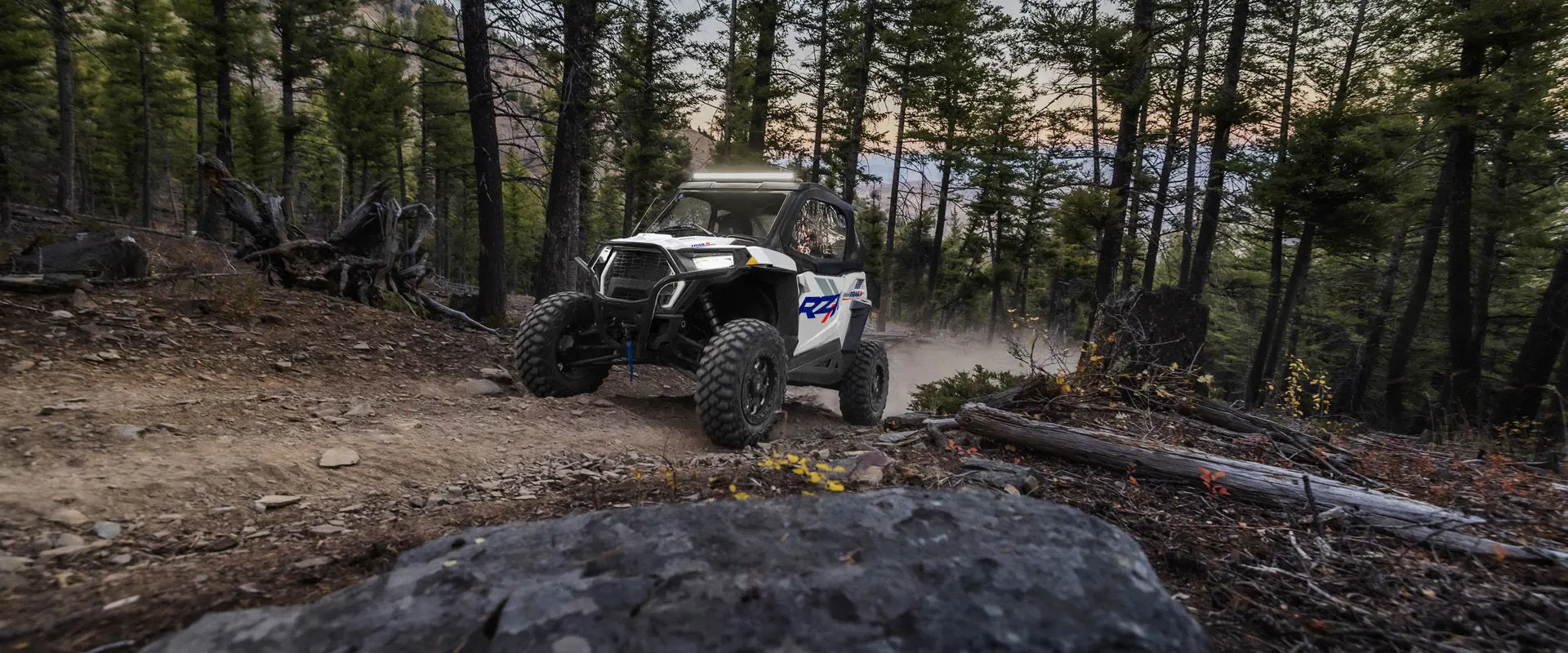Front end view of a 2027 Polaris RZR Trail S side-by-side with lights illuminated while driving through the forest.