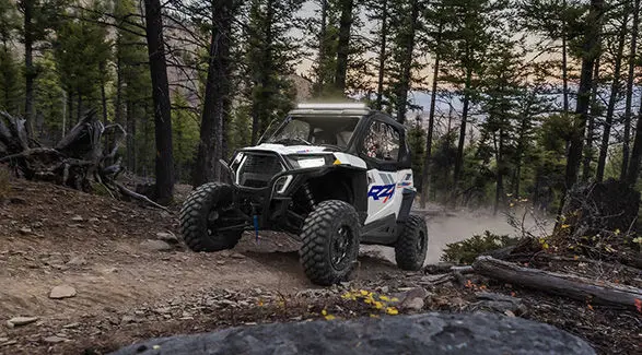 Front end view of a 2027 Polaris RZR Trail S side-by-side with lights illuminated while driving through the forest.