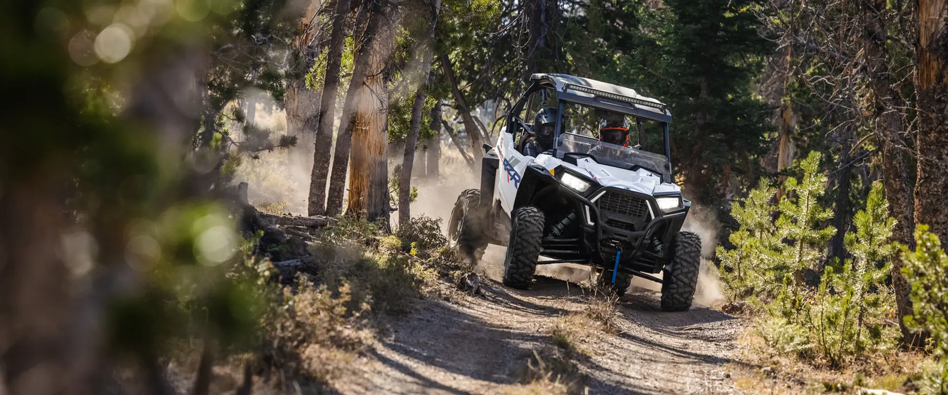 Front angle view of a 2027 Polaris RZR Trail S side-by-side driving on a tight trail in the forest.