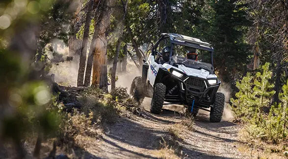 Front angle view of a 2027 Polaris RZR Trail S side-by-side driving on a tight trail in the forest.