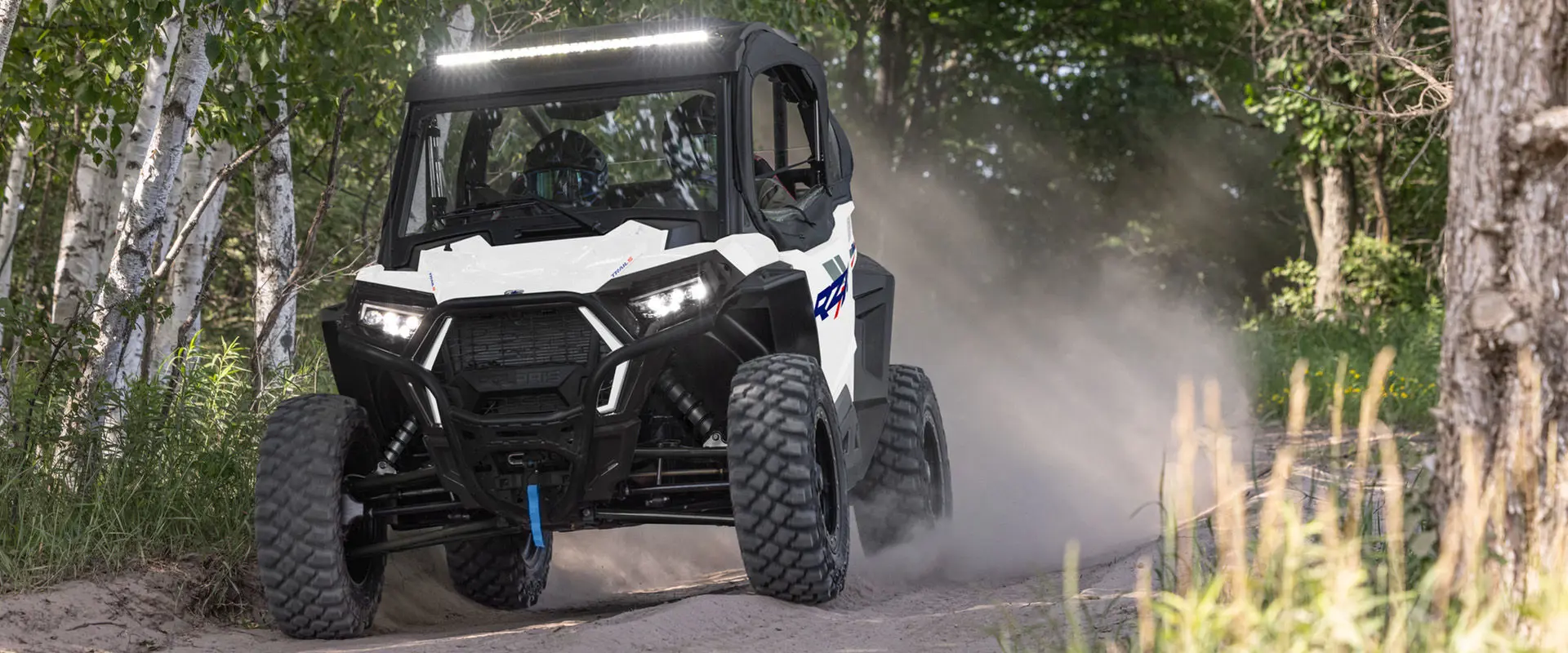 2 riders in a 2027 Polaris RZR Trail S side-by-side cruising down a tight forest trail.
