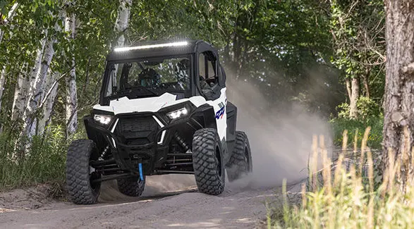 2 riders in a 2027 Polaris RZR Trail S side-by-side cruising down a tight forest trail.