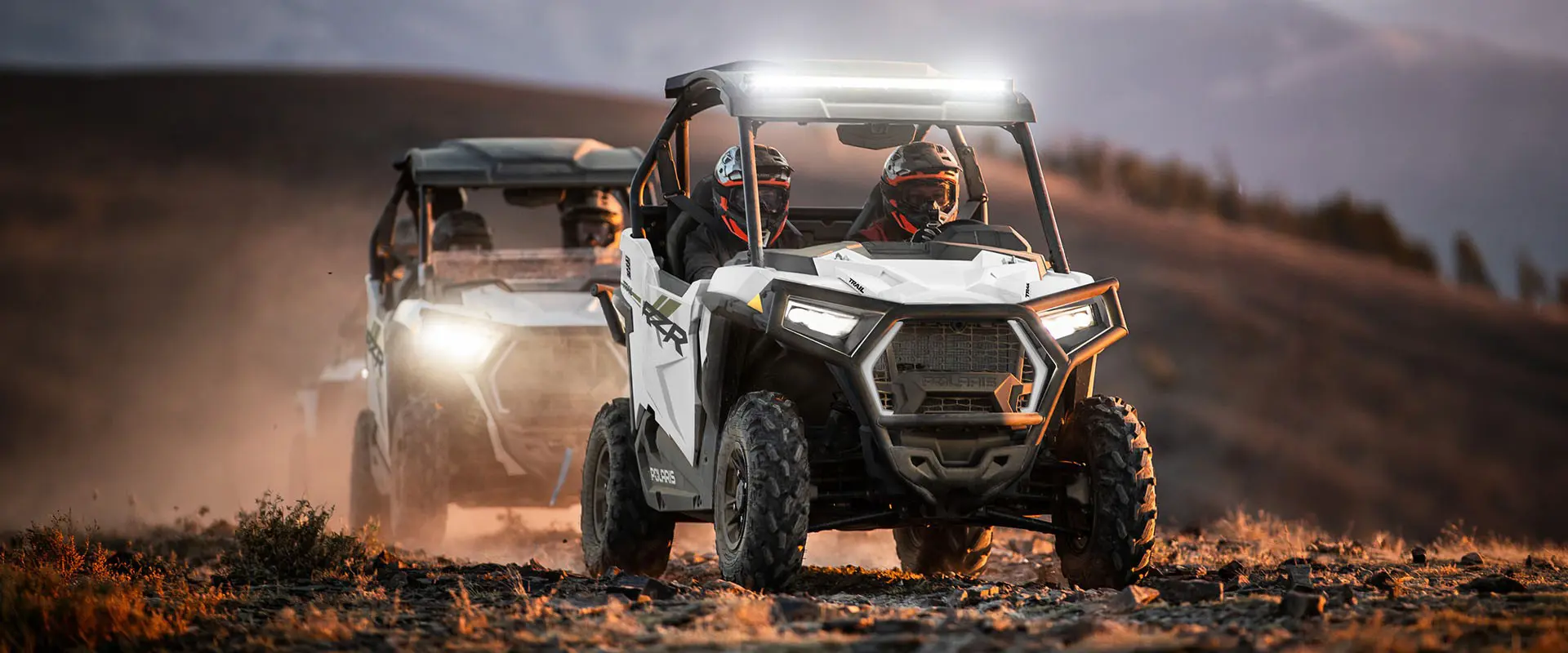 Two 2027 Polaris RZR Trail side-by-sides cruising on a trail with illuminated headlights.