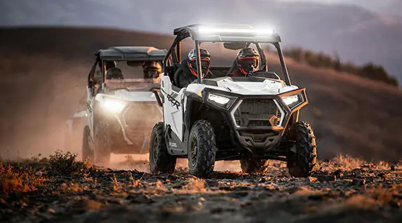 Two 2027 Polaris RZR Trail side-by-sides cruising on a trail with illuminated headlights.