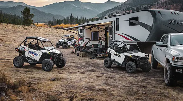 People sitting by their camper with three 2027 Polaris RZR Trail side-by-sides parked nearby.