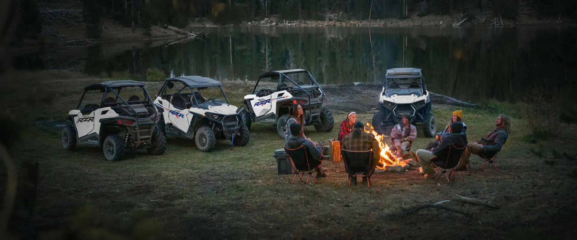 8 people around a campfire with four 2027 Polaris RZR Trail side-by-sides parked nearby.