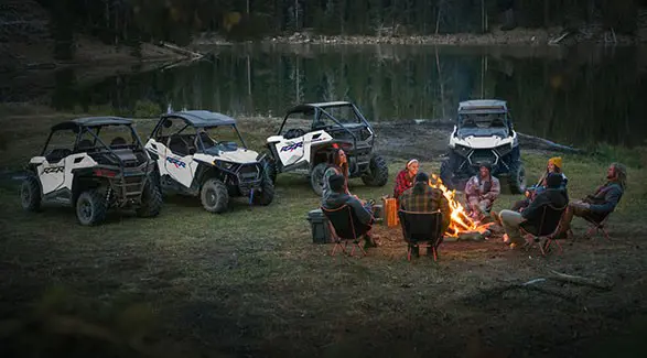 8 people around a campfire with four 2027 Polaris RZR Trail side-by-sides parked nearby.