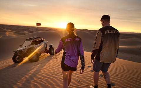 Polaris RZR Pro R SxS parked among sand dunes at sunset, with a couple wearing Polaris RZR apparel walking beside the vehicle in a desert landscape.