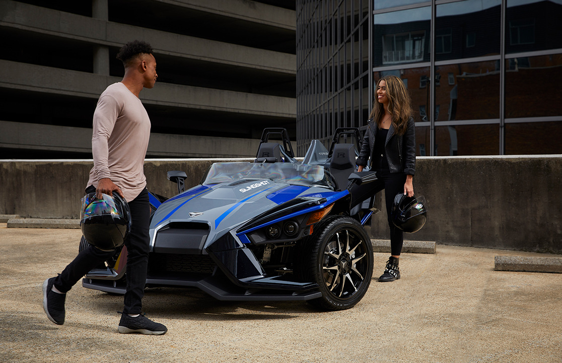 A parked Slingshot vehicle near two people with helmets on their hands