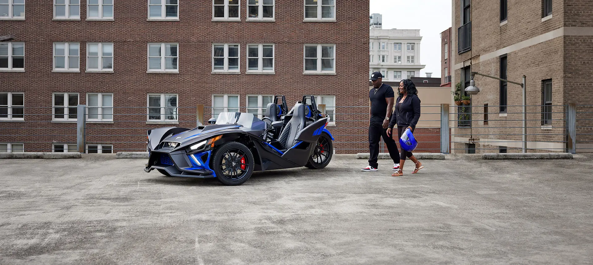 a couple walking towards a graphite blue polaris slingshot r three-wheeled roadster