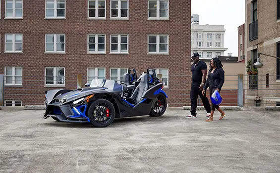 a couple walking towards a graphite blue polaris slingshot r three-wheeled roadster
